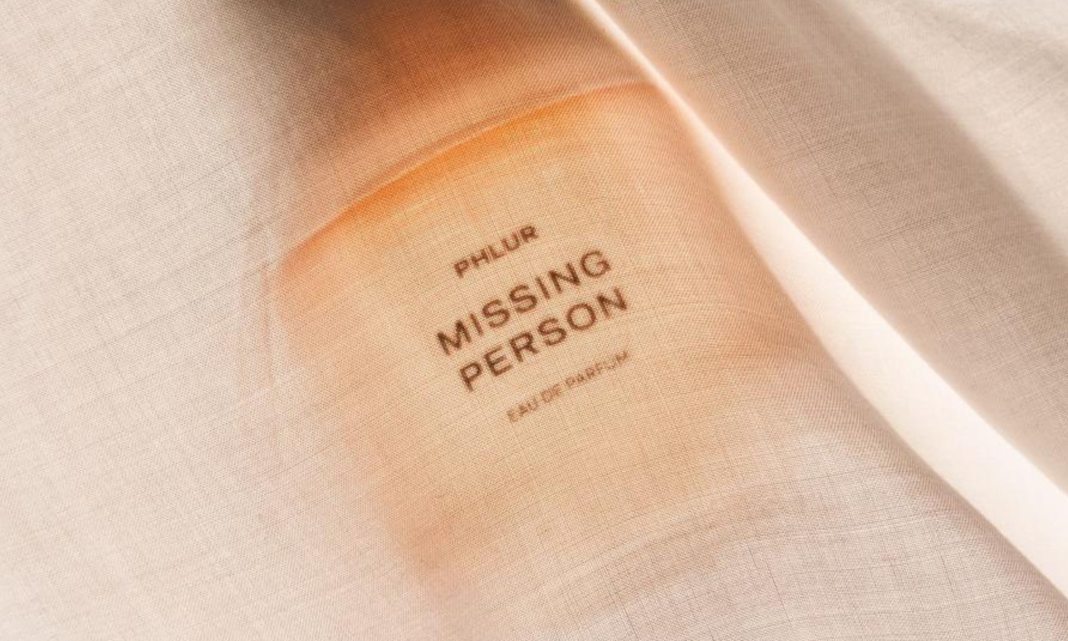 Top-down view of a Phlur Missing Person Eau de Parfum bottle showing through a sheer white linen fabric under warm light.