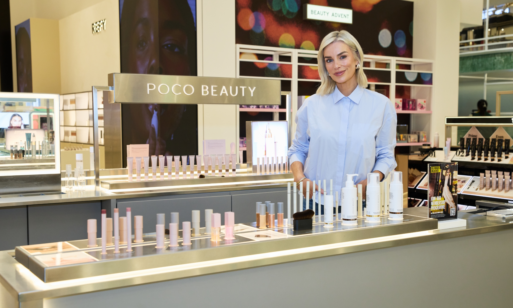 Pippa O'Connor standing behind a illuminated metal Poco Beauty display counter filled with makeup products inside Selfridges.