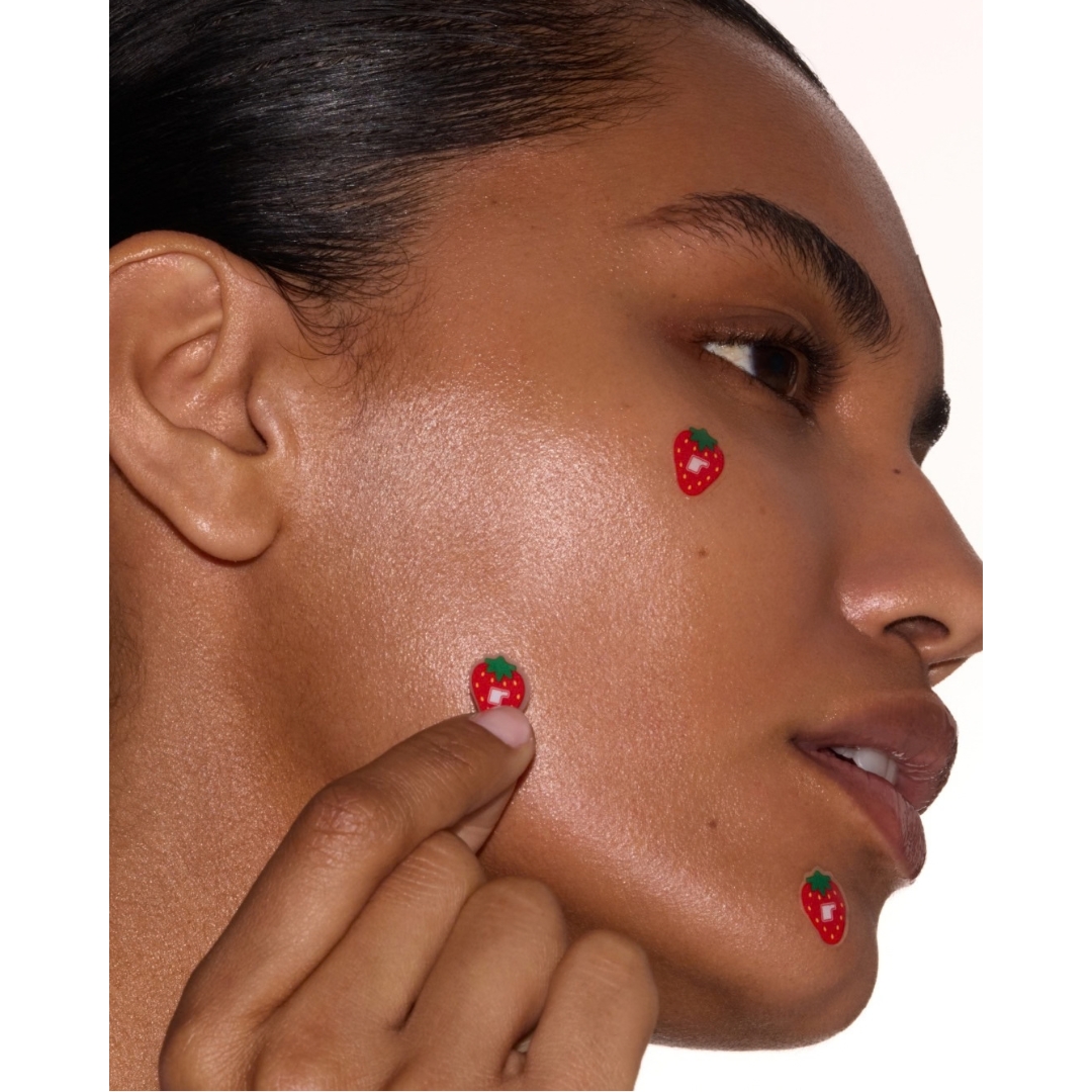 Close-up side profile of a woman applying strawberry-shaped Rhode Spotwear hydrocolloid pimple patches to her cheek.