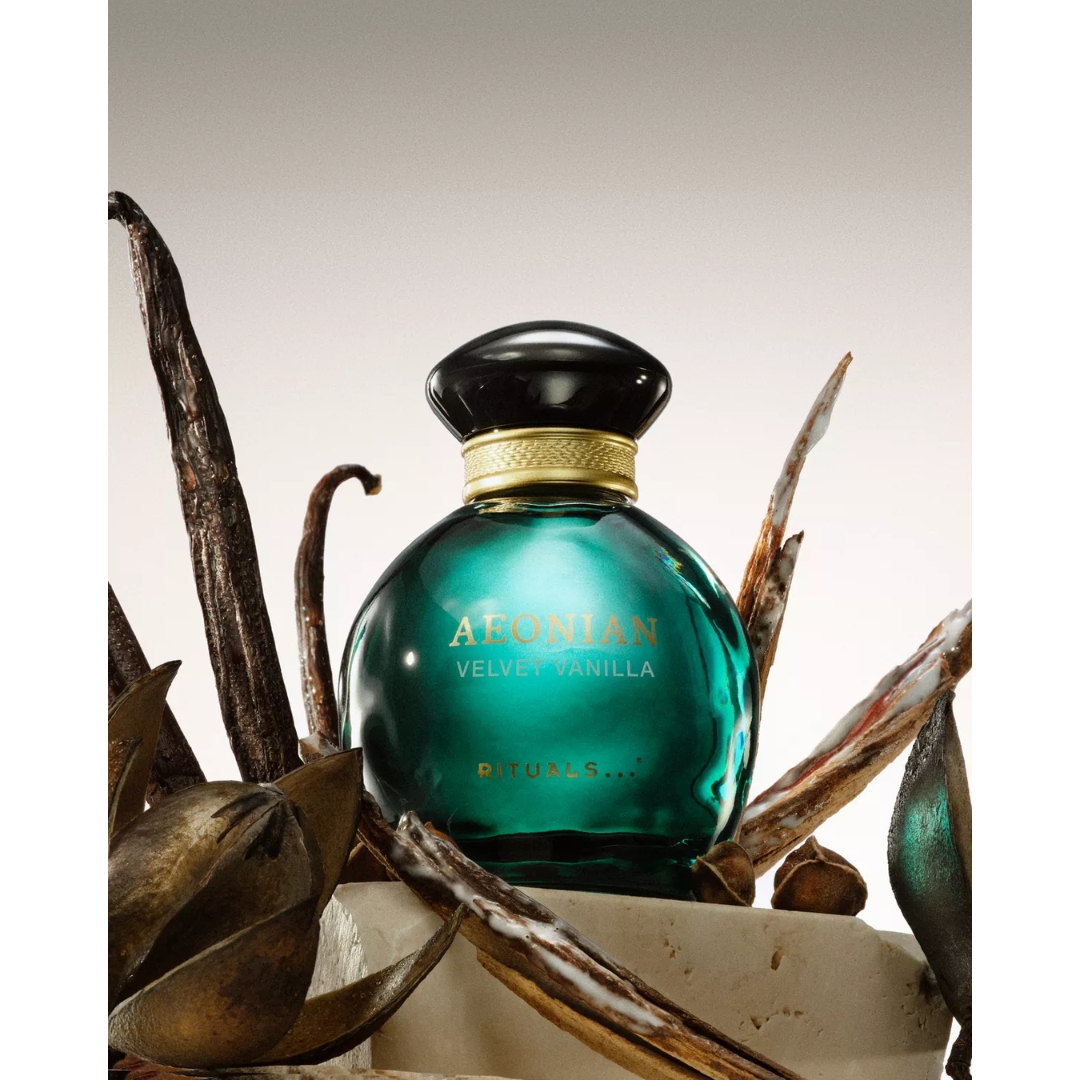 A rounded emerald green glass bottle of Rituals Aeonian Velvet Vanilla Eau de Parfum with a black cap set among vanilla pods and dried botanicals on a stone pedestal.