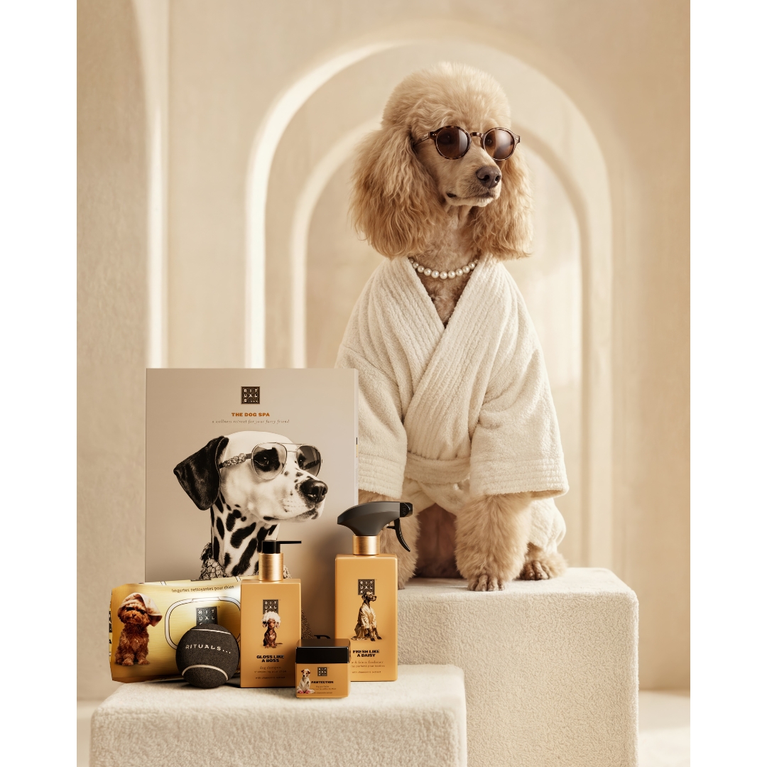 A poodle wearing a white bathrobe, sunglasses, and a pearl necklace standing beside the Rituals Dog Spa product line, including dog shampoo, grooming spray, and accessories.