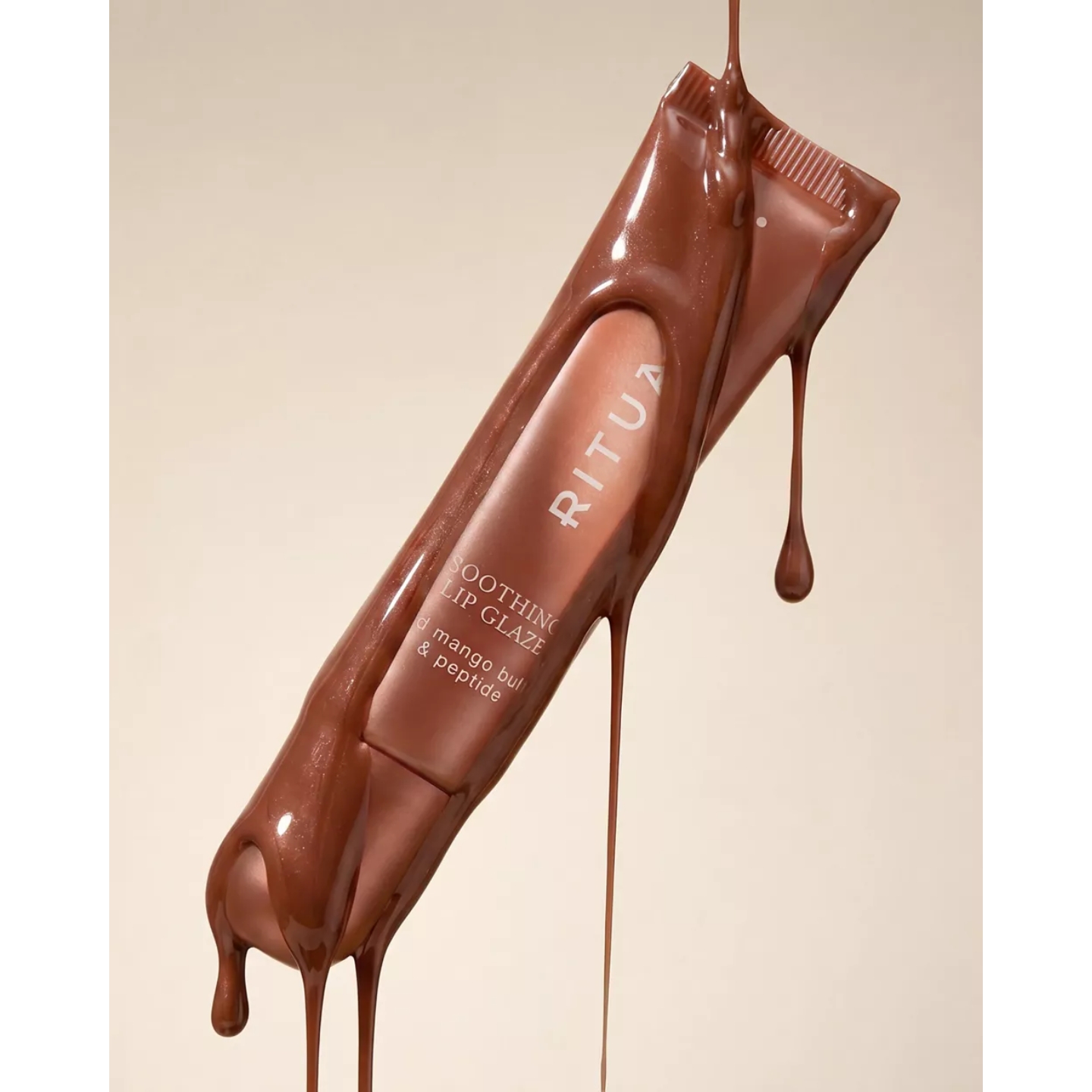 A brown tube of Rituals Soothing Lip Glaze in French Toast coated in dripping, glossy brown syrup against a warm beige background.