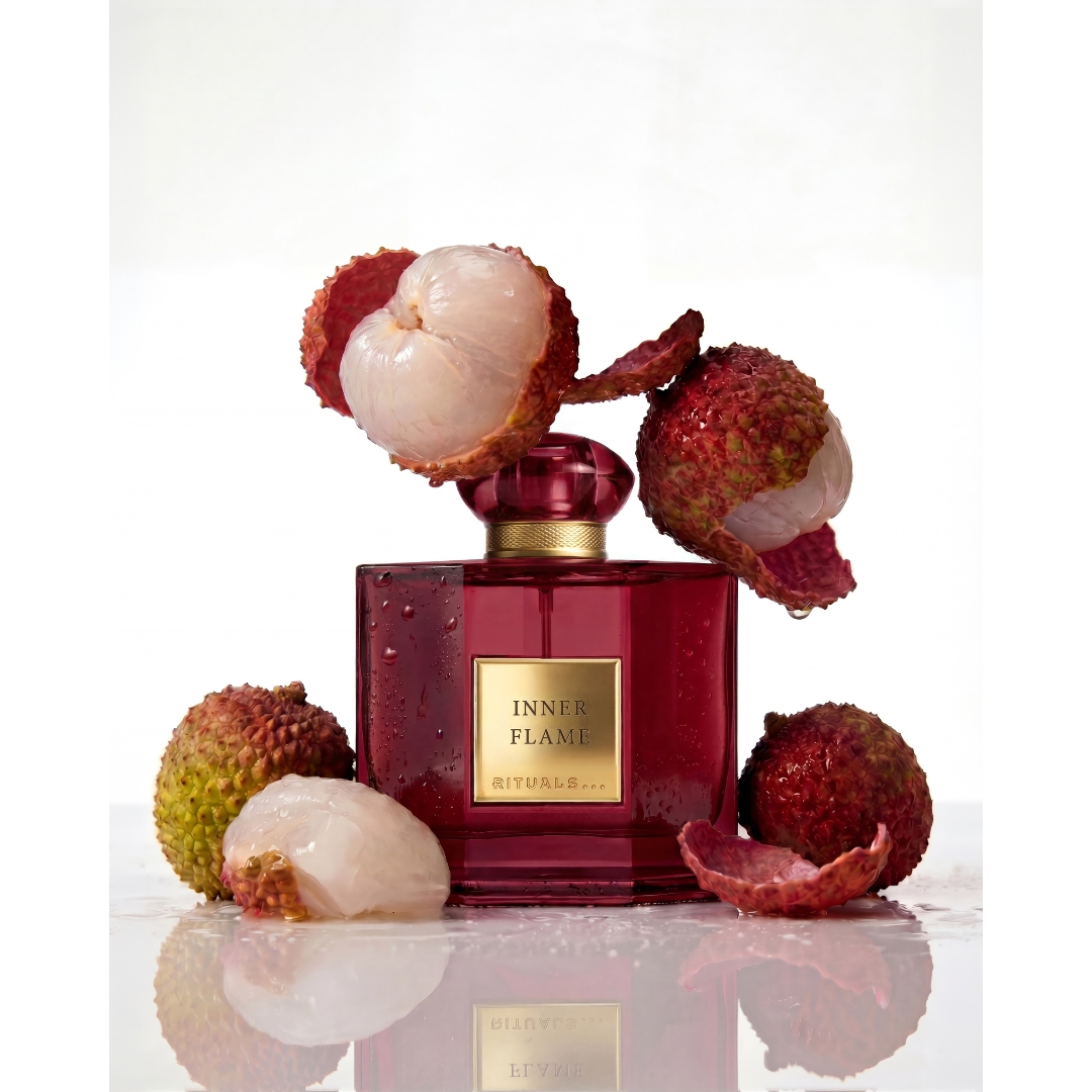 A square deep red glass bottle of Rituals Inner Flame Eau de Parfum surrounded by whole and peeled fresh lychees on a reflective white surface.