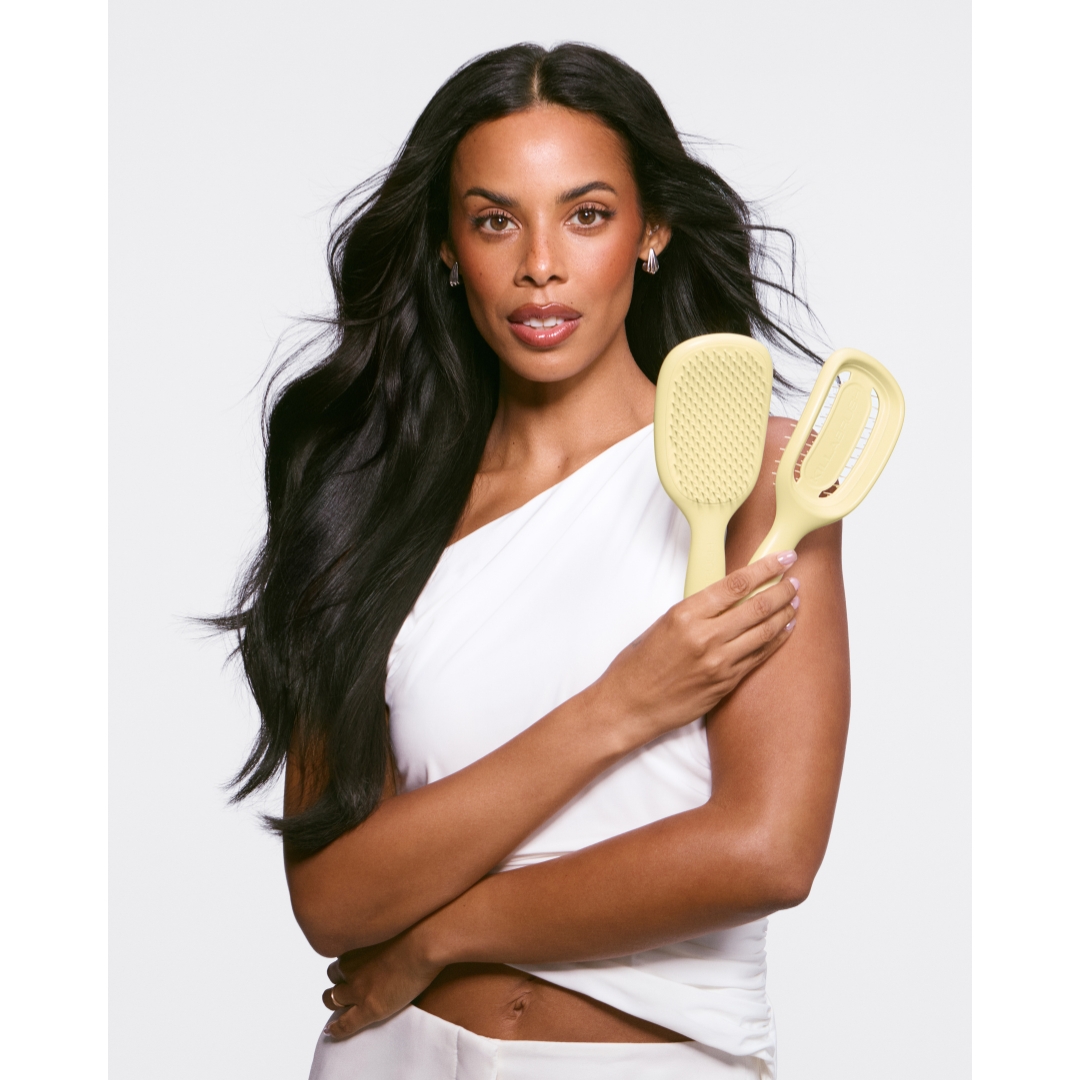 Portrait of Rochelle Humes with long dark hair wearing a white one-shoulder top, holding two pale yellow Killabrush hairbrushes.
