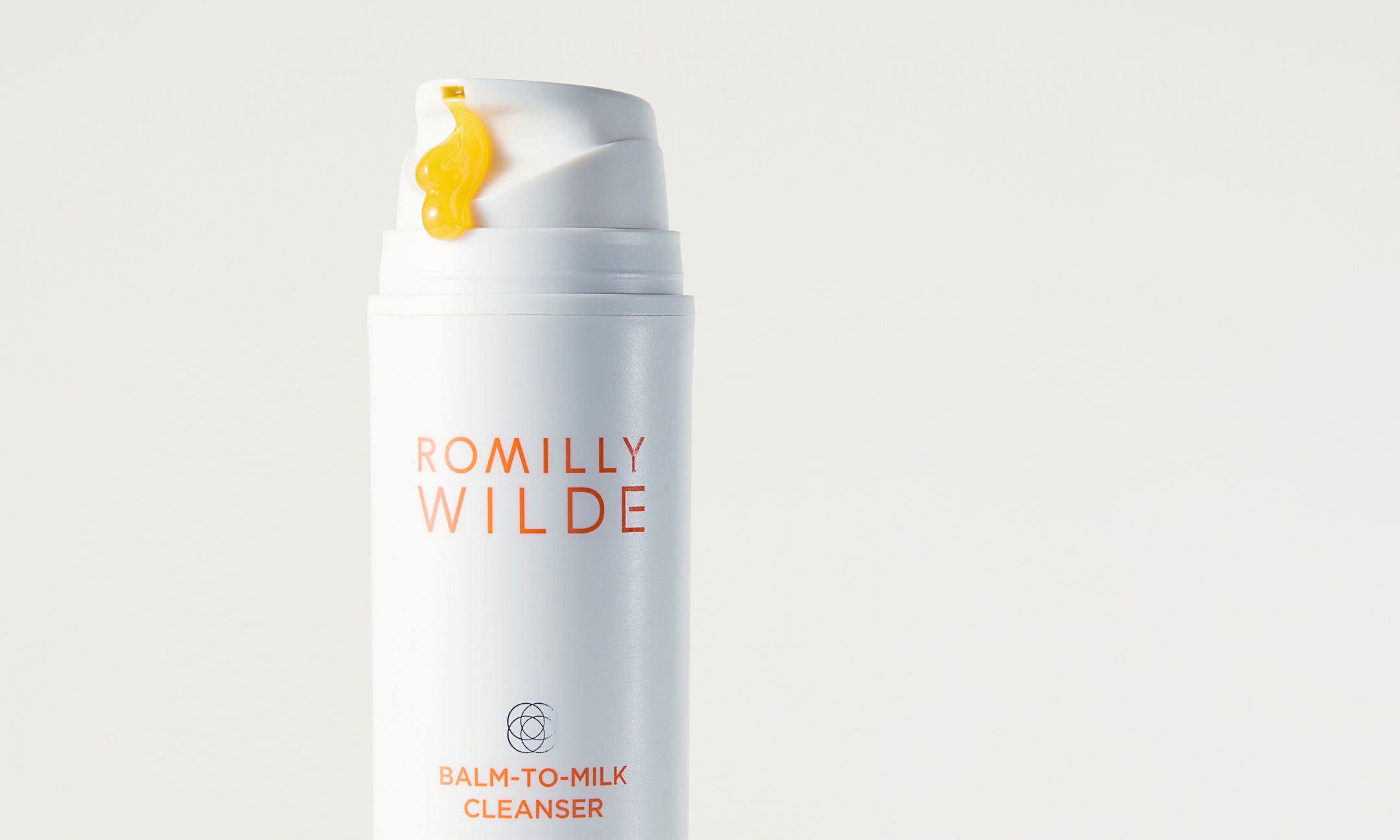 A white bottle of Romilly Wilde Balm-to-Milk Cleanser with a dollop of golden gel dispensed onto the pump top.