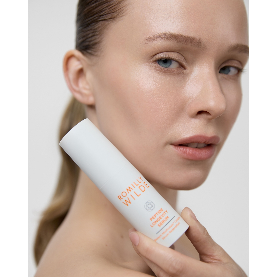 Close-up portrait of a model with slicked-back hair holding a bottle of Romilly Wilde Peptide Longevity Serum near her cheek.