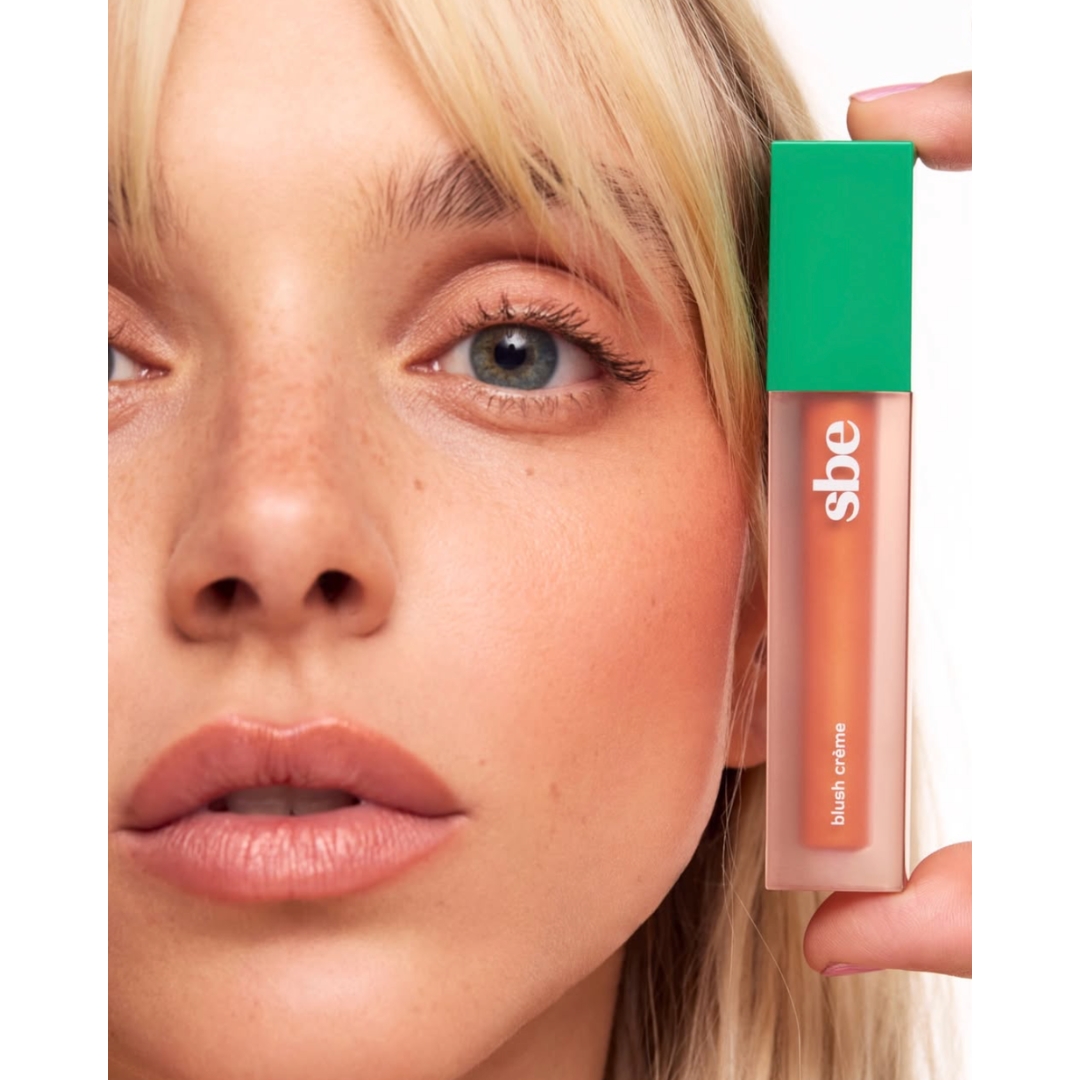Close-up portrait of a blonde model with bangs holding a tube of SBE Blush Crème with a bright green cap next to her face.