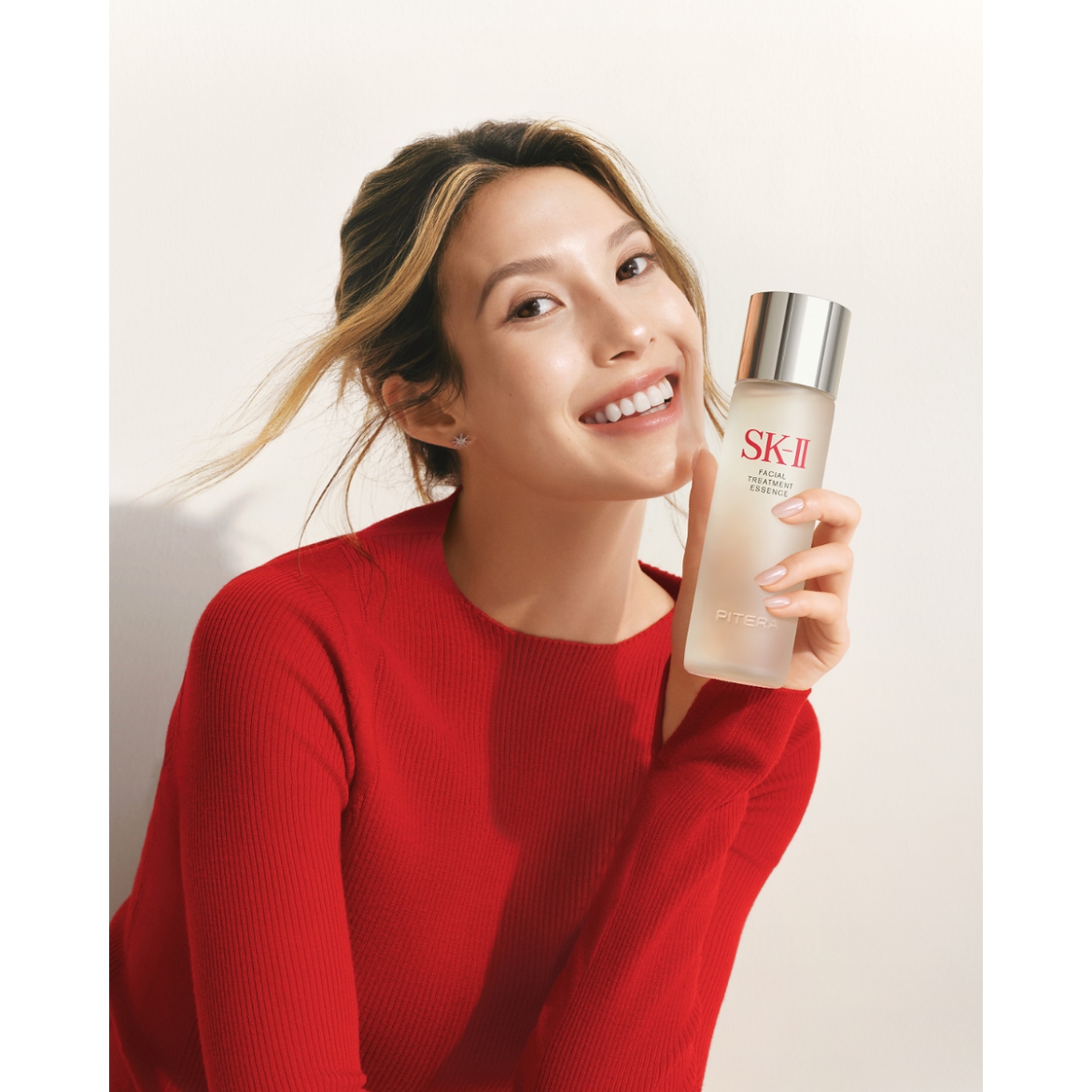Eileen Gu smiling while holding a frosted glass bottle of SK-II Facial Treatment Essence while wearing a red long-sleeve ribbed top.