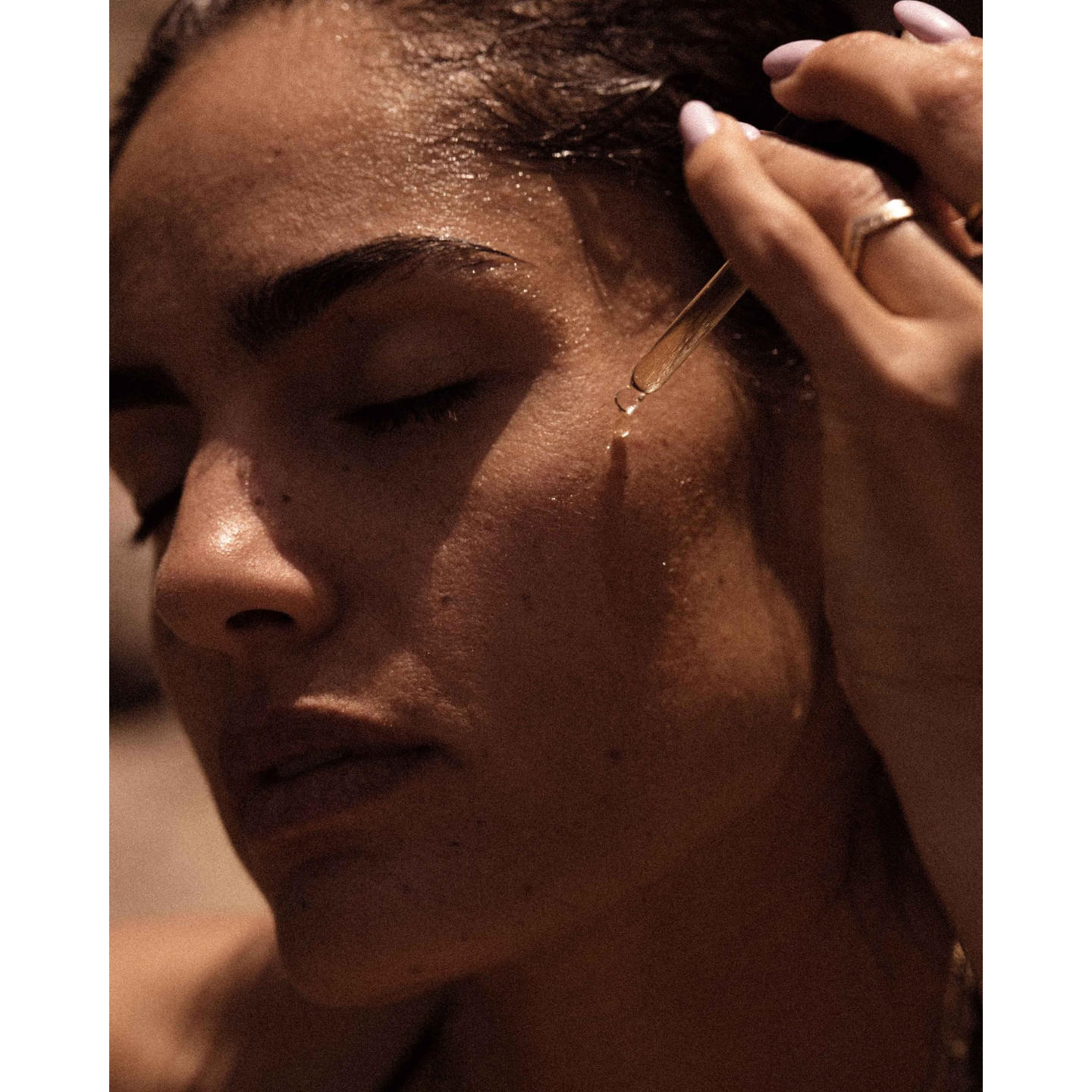 A close-up of a model applying a drop of face oil from a dropper onto her cheek, featured in the Sacra skincare UK launch announcement on Beauty News Daily.