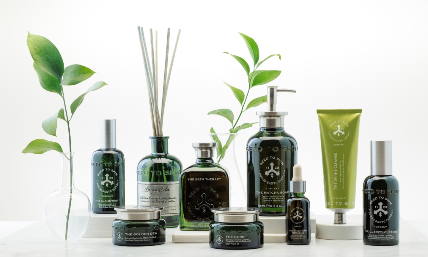 An assortment of Seed to Skin Tuscany luxury skincare and bodycare products, including dark green glass bottles, face creams, and a room diffuser, arranged among green botanical sprigs.