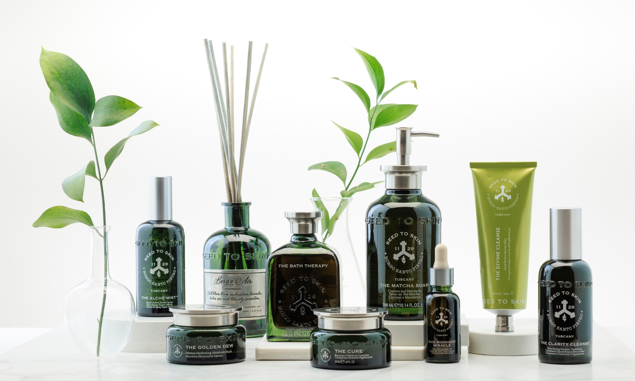 An assortment of Seed to Skin Tuscany luxury skincare and bodycare products, including dark green glass bottles, face creams, and a room diffuser, arranged among green botanical sprigs.