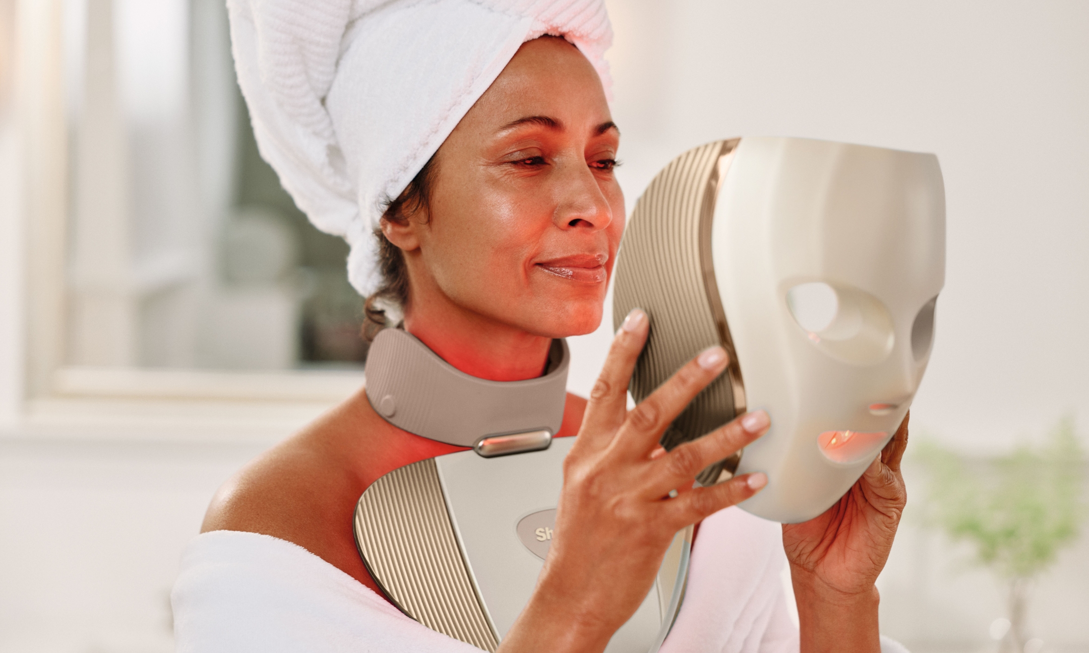 A woman wearing a white bathrobe and towel turban holding up the Shark Beauty CryoGlow LED face mask while wearing the red light therapy neck and chest attachment.