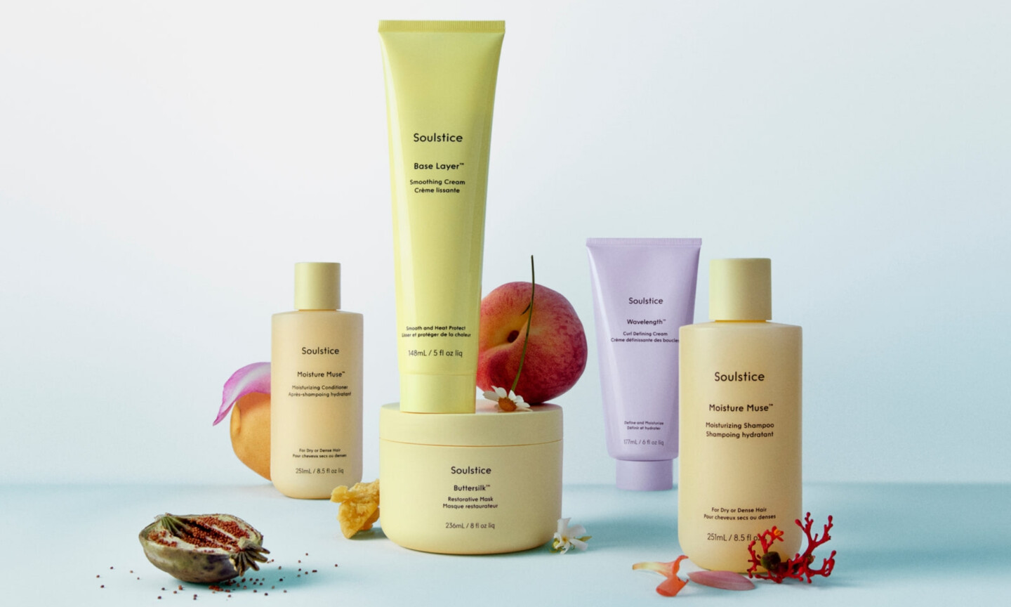 An assortment of pale yellow and lavender Soulstice hair products, including Moisture Muse shampoo and conditioner, Buttersilk mask, Base Layer smoothing cream, and Wavelength curl cream, styled on a light blue background with fresh peach slices, flower petals, and seed pods.