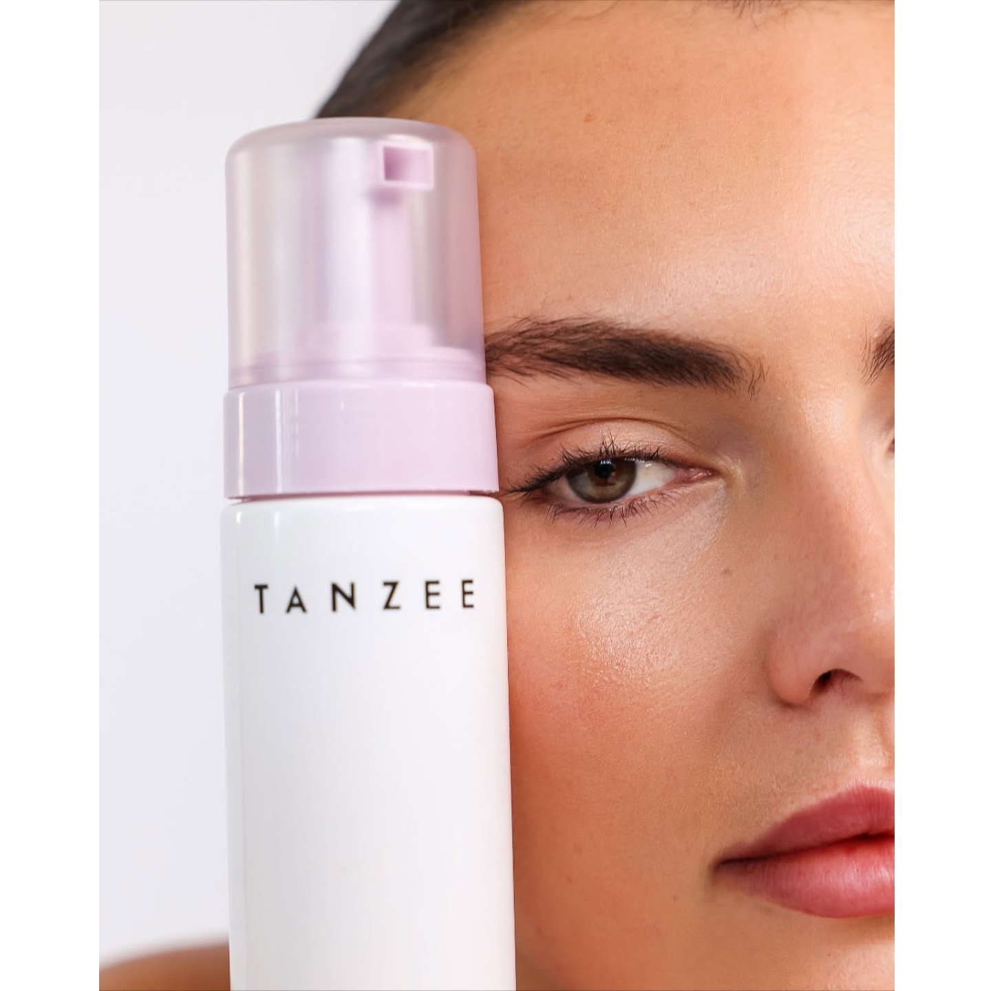 Close-up of a model holding a white Tanzee self-tanning mousse bottle with a light purple cap next to her face.