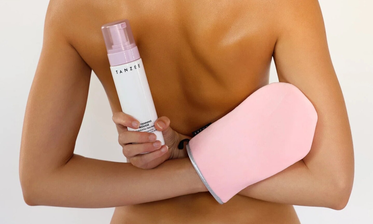Rear view of a model reaching behind her back while holding a Tanzee Self-Tanning Mousse bottle in one hand and wearing a pink tanning mitt on the other against a white background.