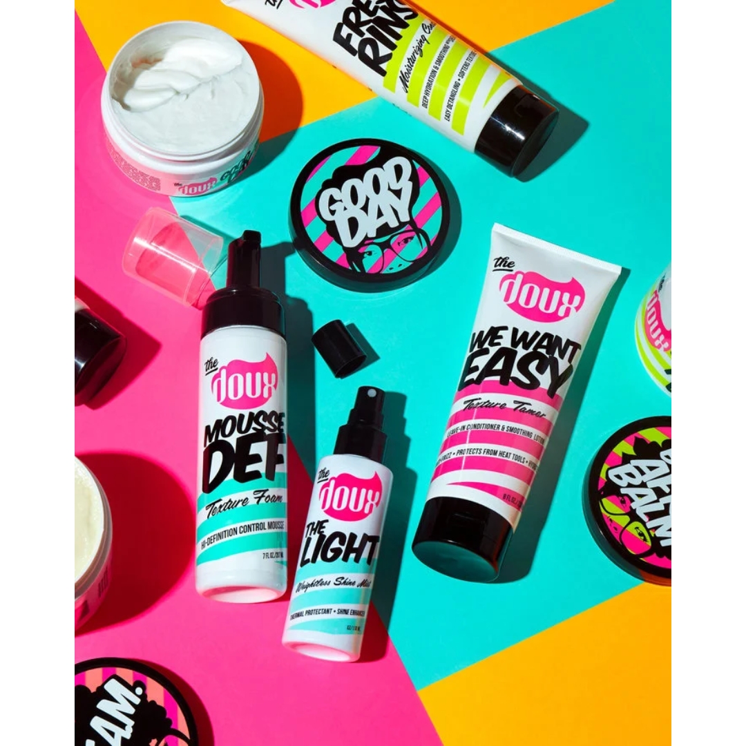A colorful flat-lay view of various The Doux haircare products, including Mousse Def Texture Foam, sprays, and jars arranged on a bright pink, yellow, and blue geometric background.