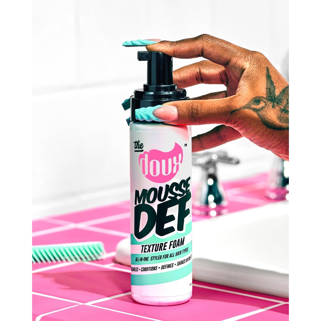A hand with mint-green nails and a bird tattoo pressing the pump of a bottle of The Doux Mousse Def Texture Foam on a pink-tiled bathroom counter.