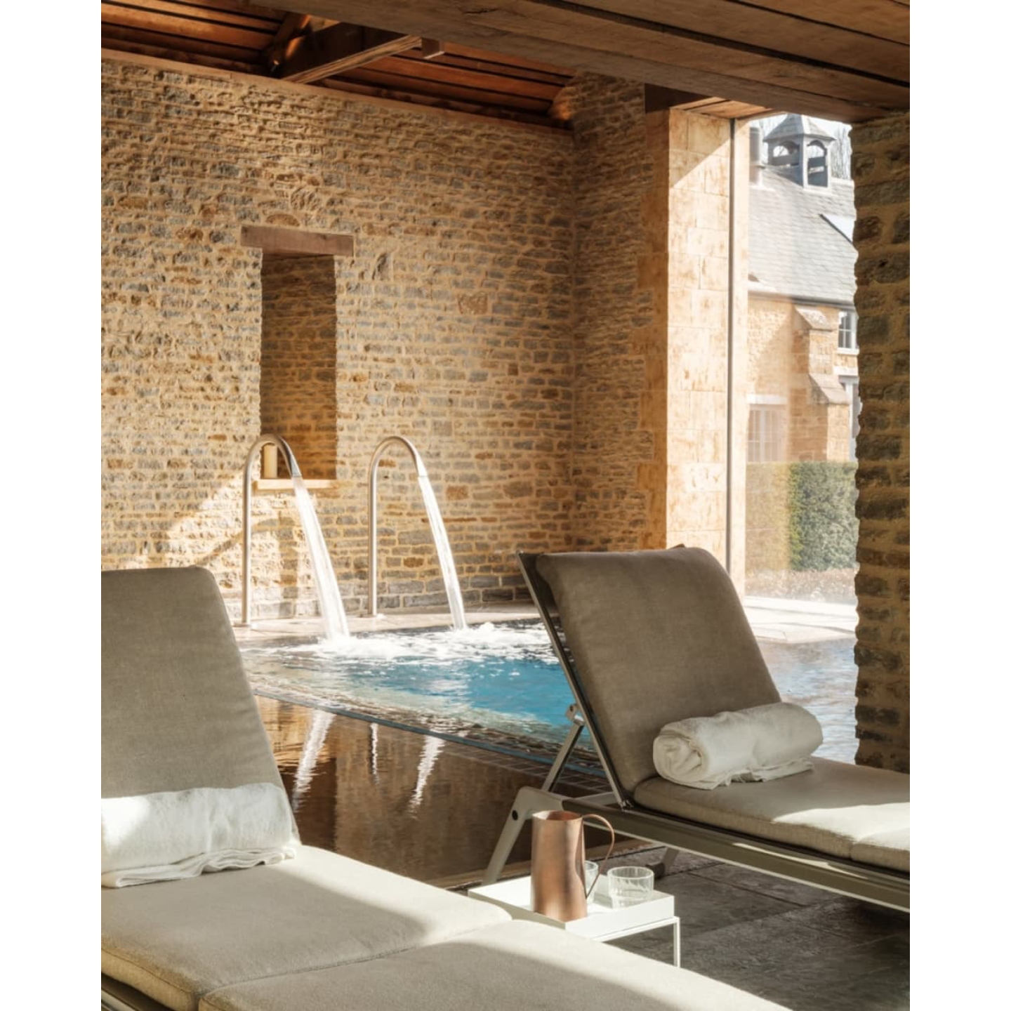 Two neutral-toned lounge chairs beside an indoor-outdoor hydrotherapy pool with cascading water fountains, set against exposed Hadspen limestone walls at The Newt in Somerset spa.
