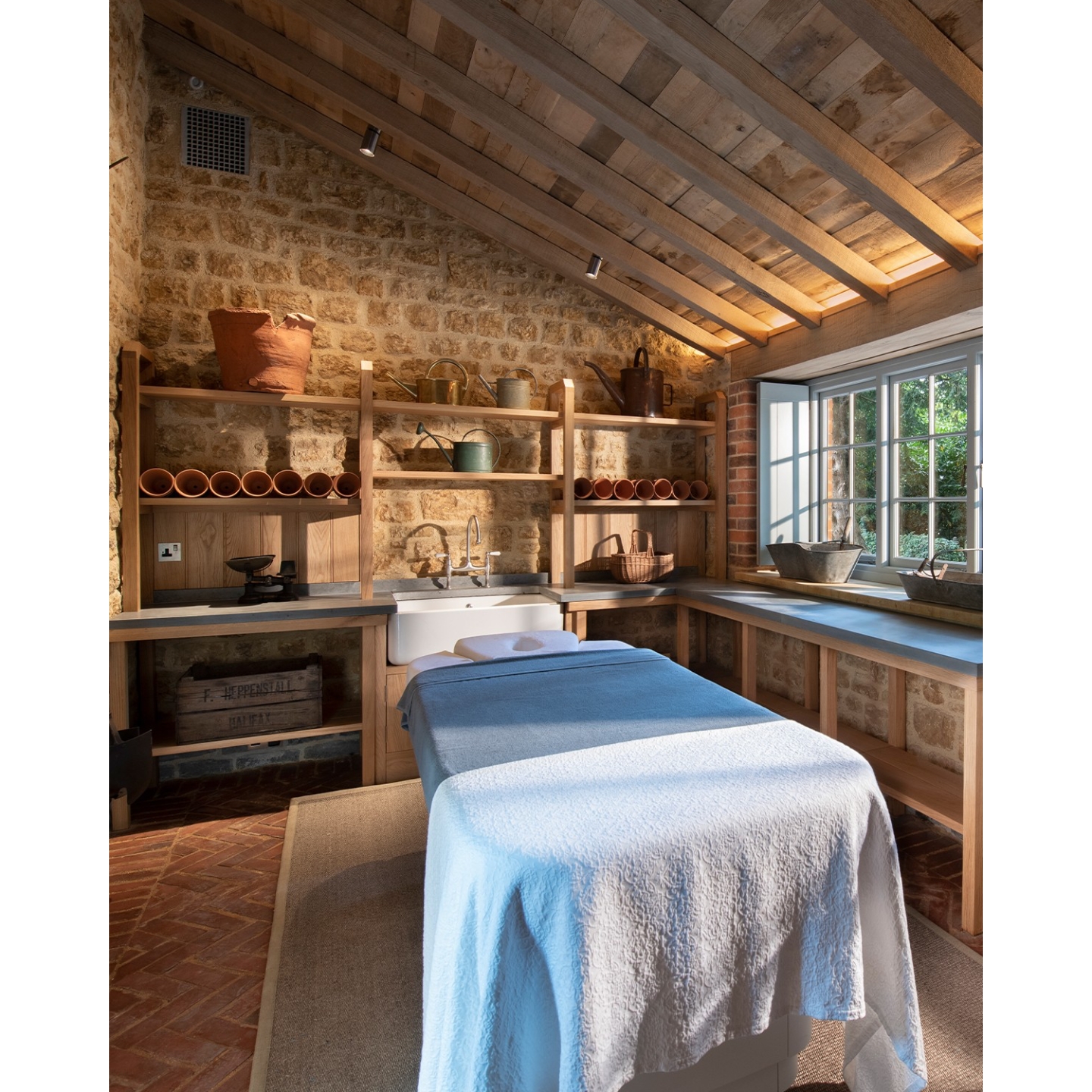 A massage table draped in white linens inside a rustic treatment room at The Newt Spa, featuring stone walls, wooden beams, terracotta pots, and natural light from a large window.