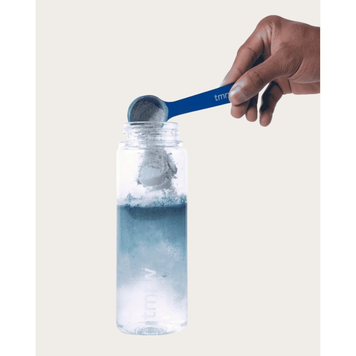A hand using a blue scoop to pour blue Tmrw Daily supplement powder into a clear water bottle against a light beige background.