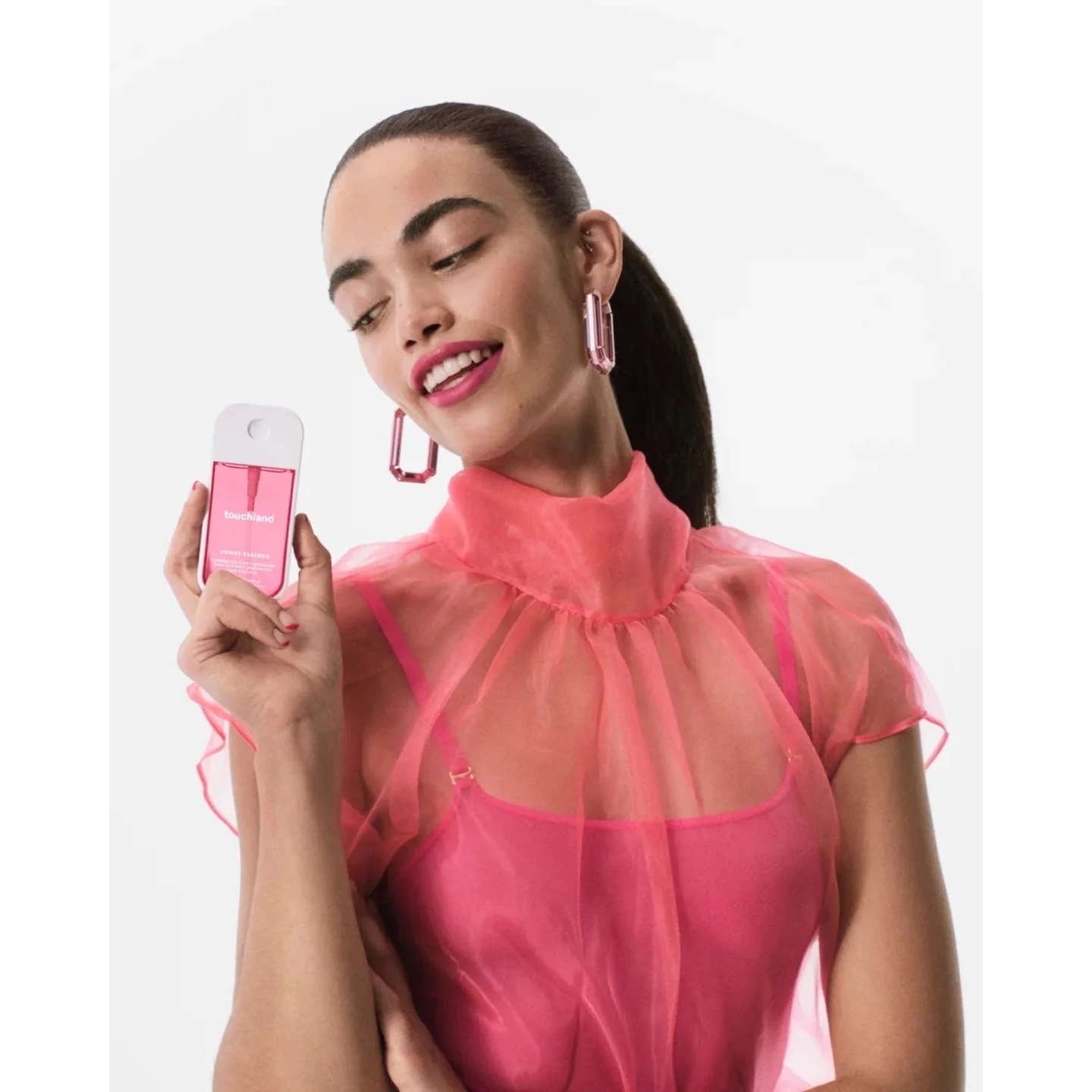 A model wearing a bright pink sheer top and pink statement earrings holding a flat pink Touchland Lush Tropicale mist spray bottle against a plain white background.