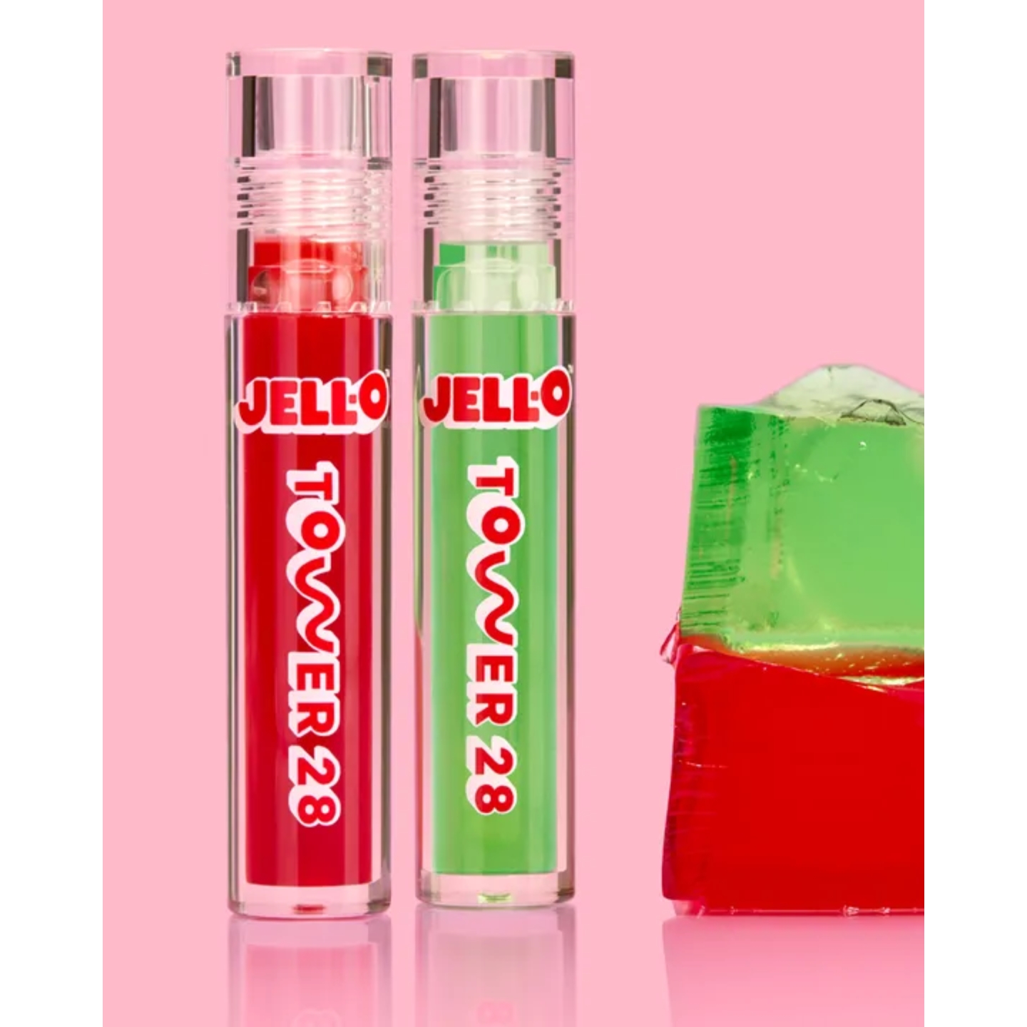 Two clear tubes of Tower 28 x Jell-O lip jelly in red and green shades standing beside stacked red and green gelatin cubes against a pink background.