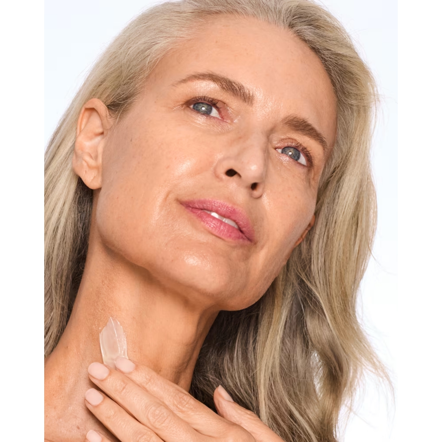 Close-up of a blonde model applying a swatch of Trinny London The Elevator neck and décolleté cream to her neck against a plain white studio background.