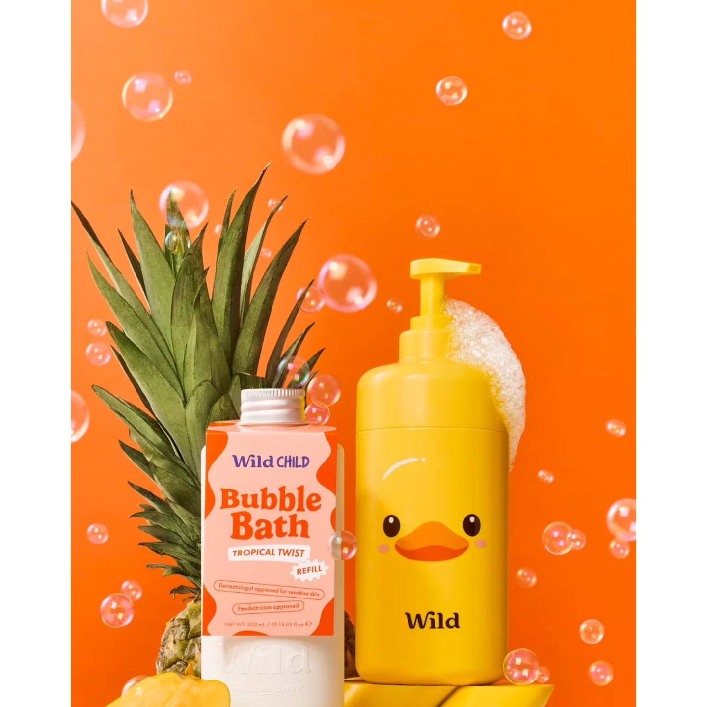 A yellow rubber duck-printed Wild pump bottle with soap suds next to a WildChild Tropical Twist Bubble Bath refill, set against a pineapple and floating bubbles on a vibrant orange background.