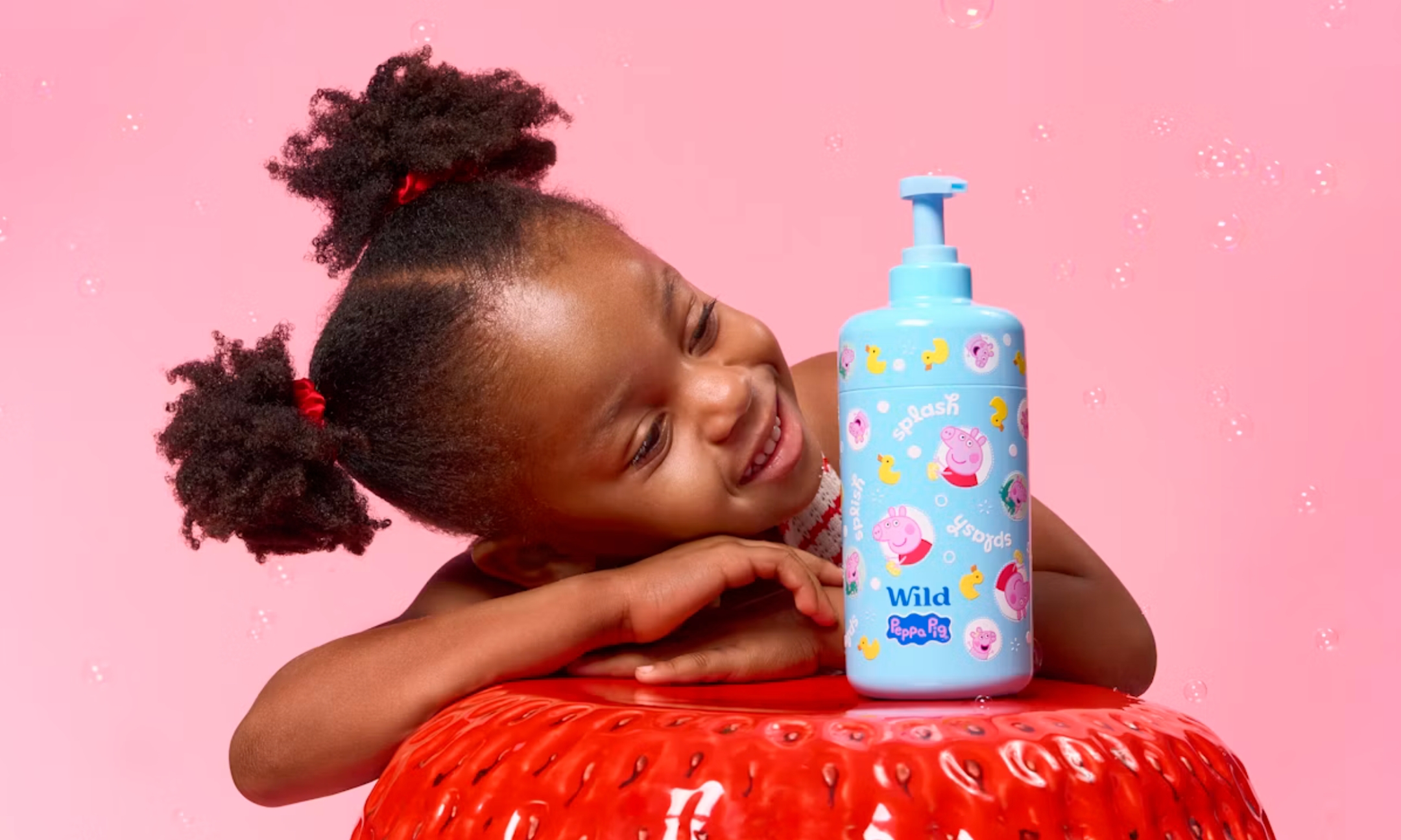 A young girl resting her head on a large red strawberry prop beside a blue Wild Peppa Pig pump bottle with floating bubbles against a pink background.