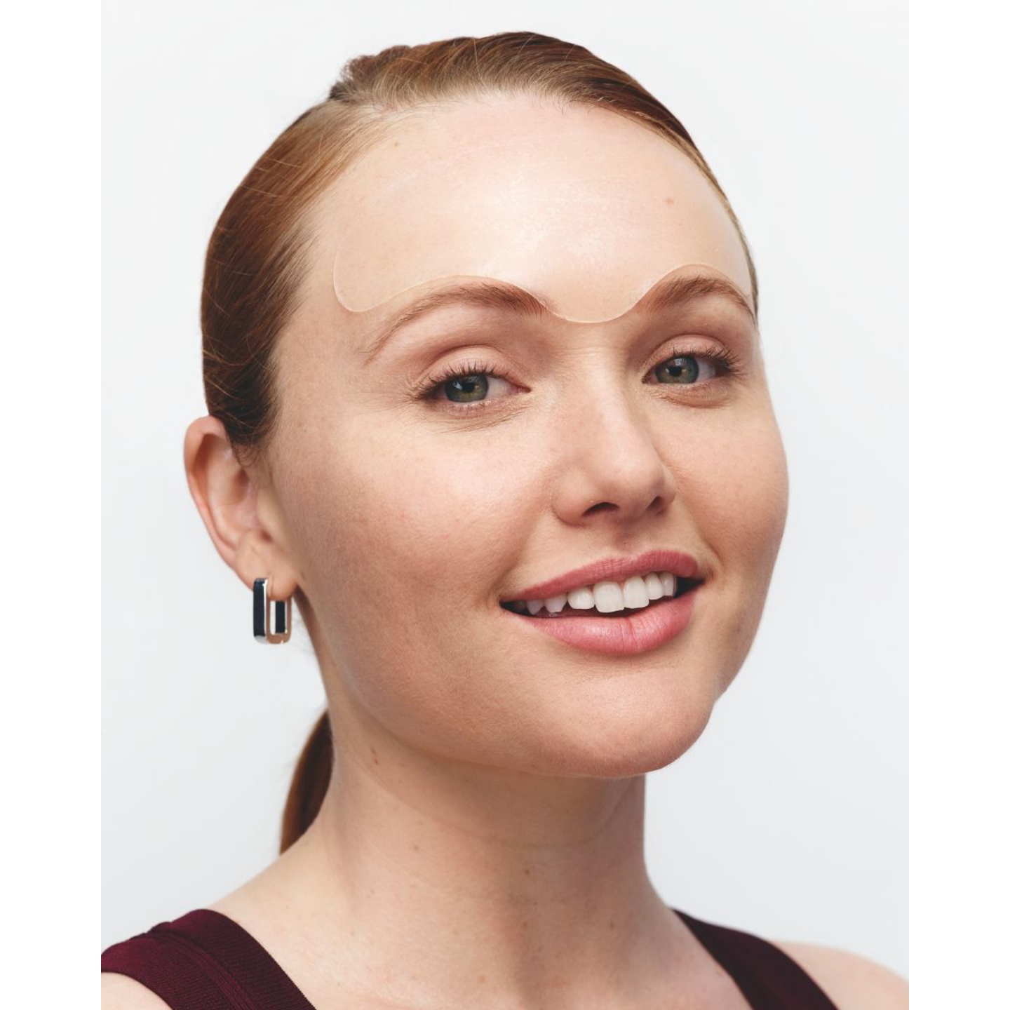 Model smiling with a clear Wrinkles Schminkles silicone forehead patch applied above her eyebrows against a plain white background.