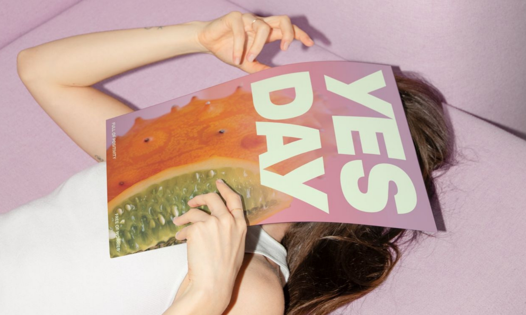 A model lying down on a light purple couch holding a Yes Day skincare poster with a kiwano melon illustration over her face, featured in the brand's Urban Outfitters launch announcement on Beauty News Daily.