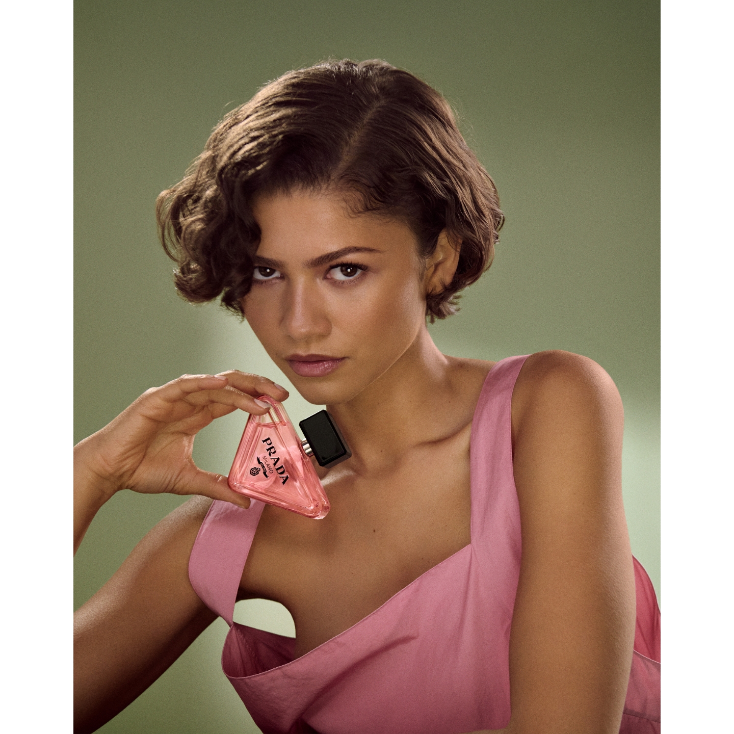 Campaign portrait of Zendaya wearing a pink top against a soft green background, holding a triangular pink-purple Prada Paradoxe Sweet Chemistry perfume bottle up to her neck.