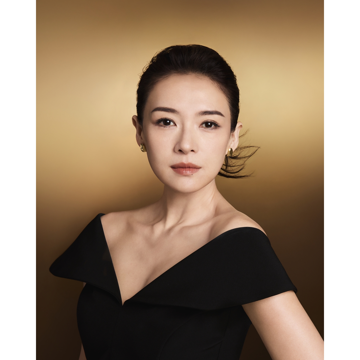 Actress Zhang Ziyi in an off-the-shoulder black dress with sleek updo hair in a campaign image for Clarins Precious skincare against a warm gold background.
