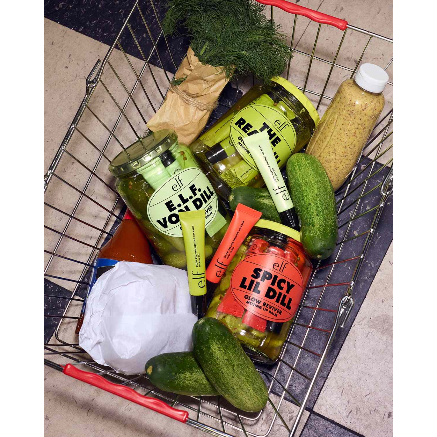 A wire shopping basket filled with e.l.f. Glow Reviver Melting Lip Balms in pickle-themed jars labelled The Real Dill, e.l.f. von Dill, and Spicy Lil’ Dill alongside fresh cucumbers, dill sprigs, and hot sauce.